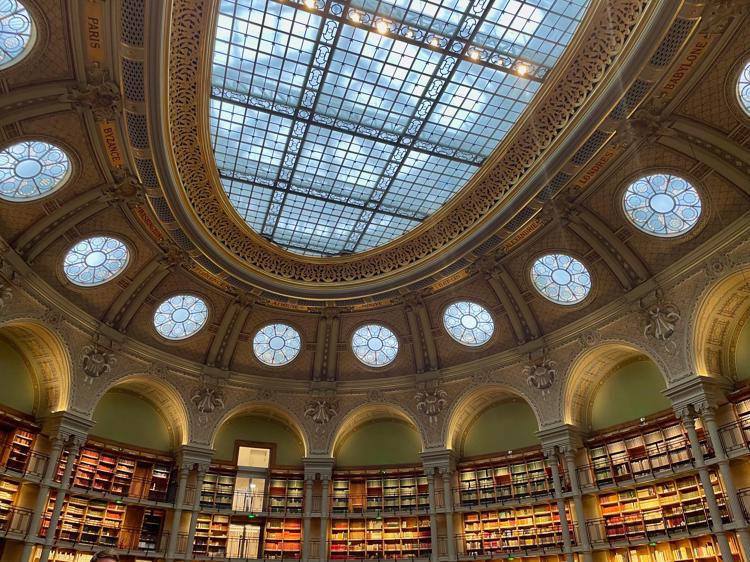 Looking up towards the ceiling. There's a large oval window at the very top of the ceiling, then smaller circular windows around it. Above each window is the name of a city; below it is an archway with bookshelves filling the archway. Each archway is wide, enough to hold six bookshelves.